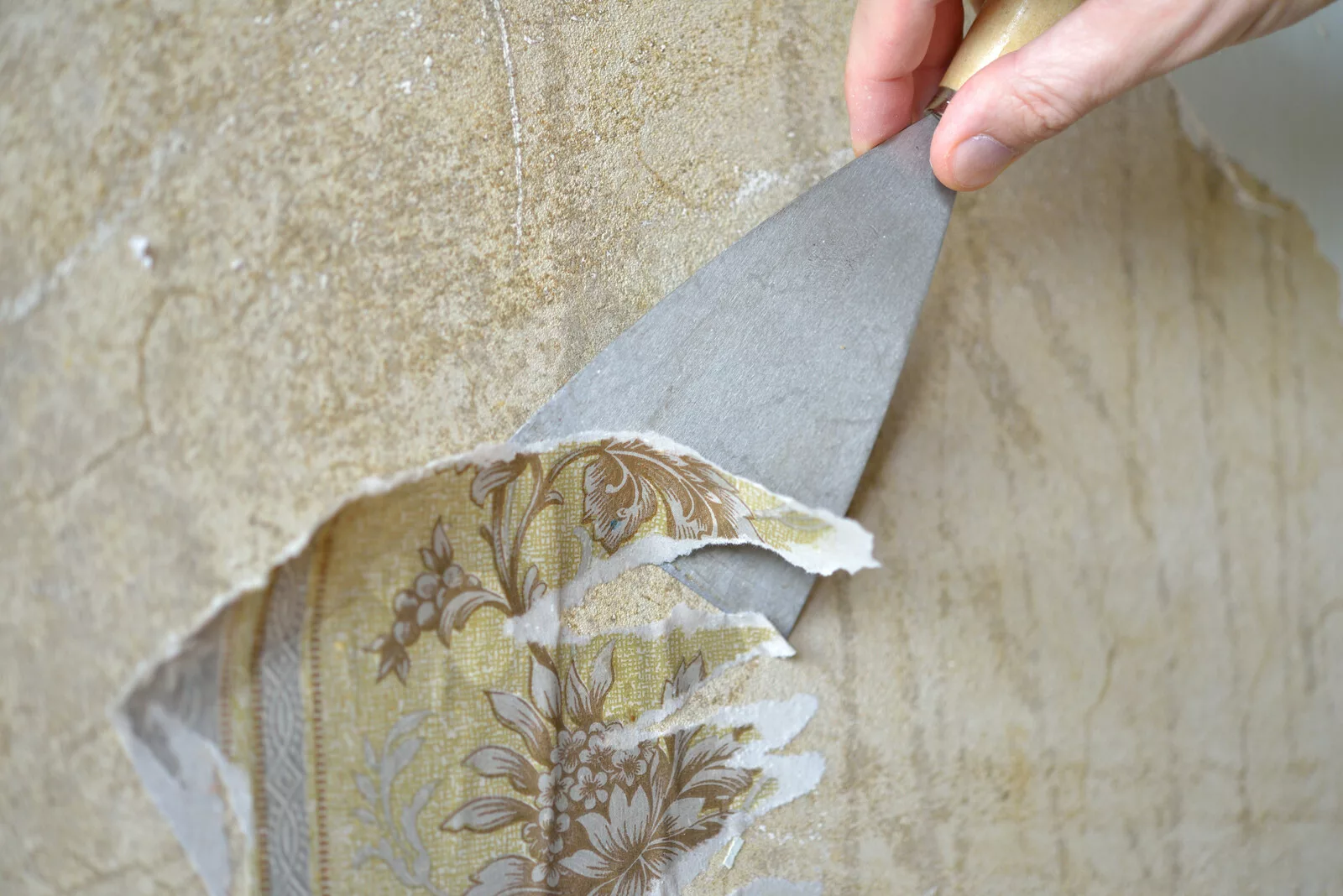 Hand using a scraper to remove old floral wallpaper during home renovation.