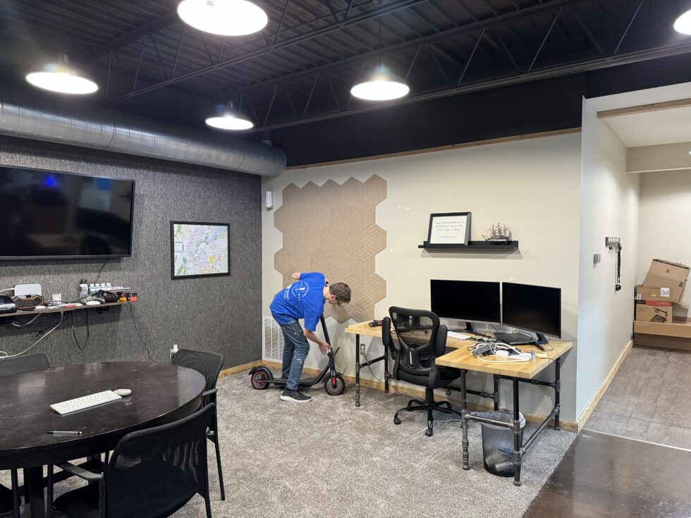 Young man assembling an electric scooter in a modern office space with desks and a conference table.