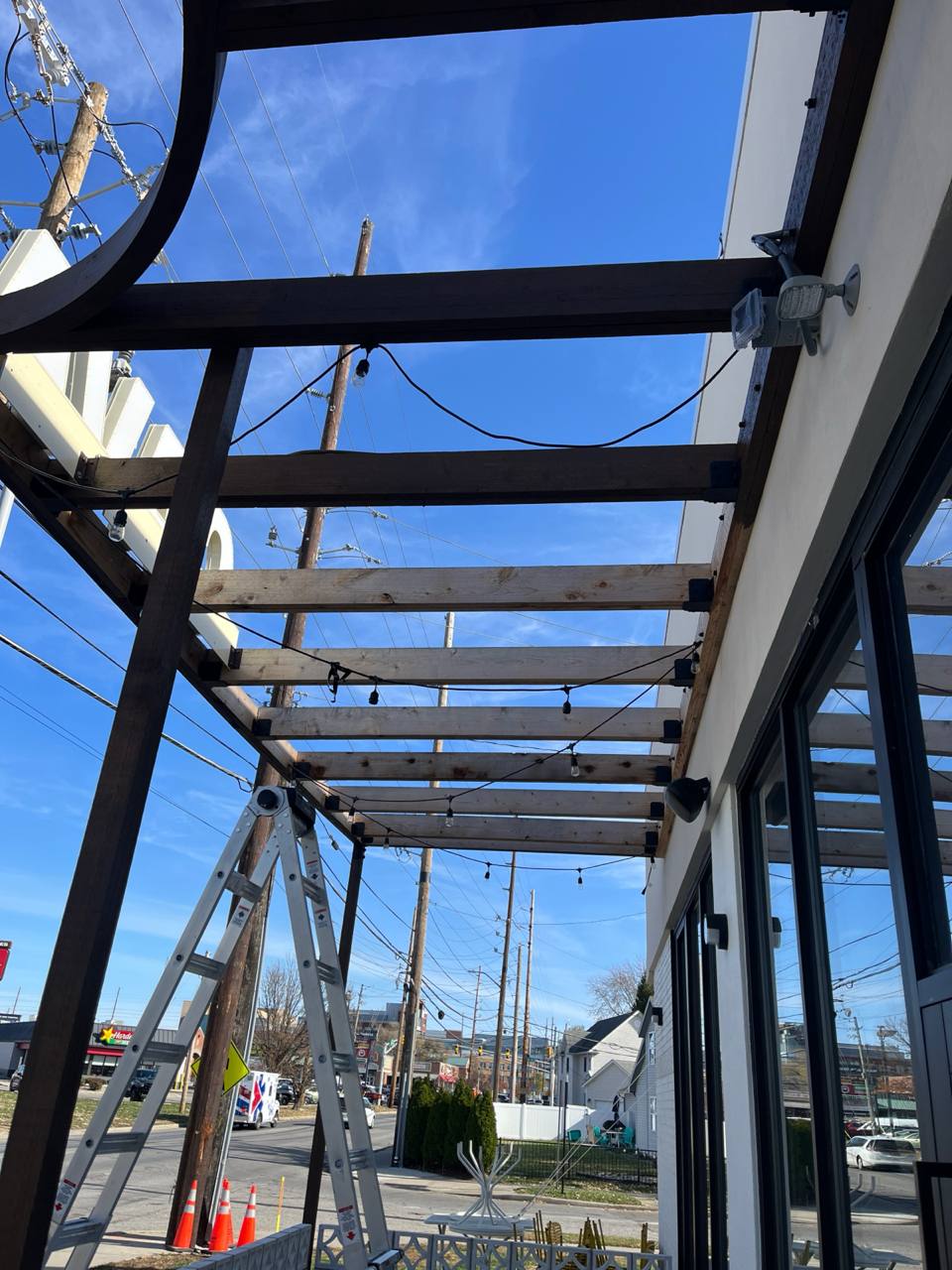 Pergola Construction Outdoor Lighting Installation Cava canopy exterior staining: wooden beams and string lights under a blue sky.