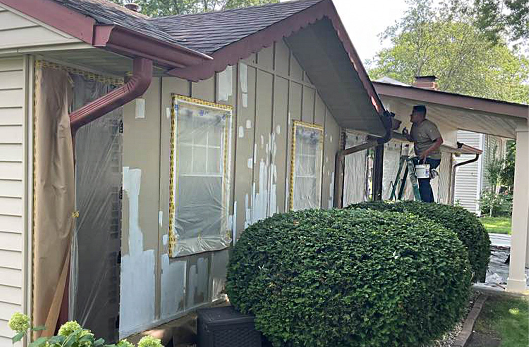 Home exterior prep for painting: masking windows, covering bushes, and painter on ladder.